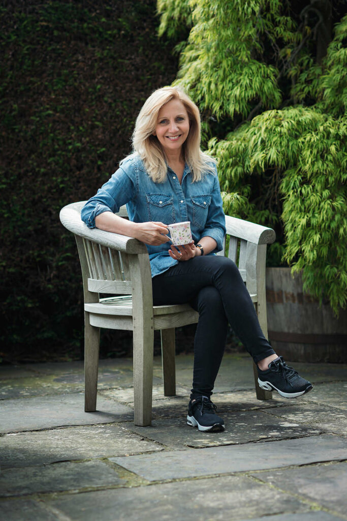 Hilary Robinson sitting on a wooden garden chair, holding a cup of tea.
